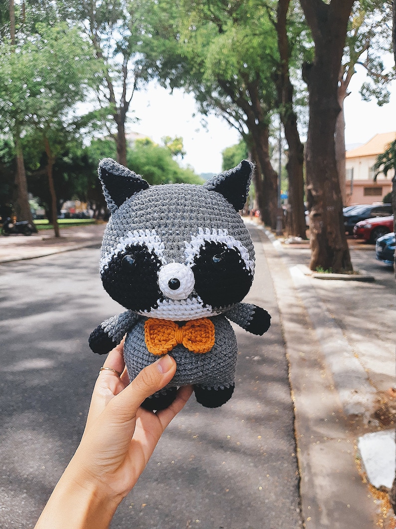May include: A crocheted raccoon plush toy wearing a bow tie. The toy is grey with black eyes and a white nose. The bow tie is orange.