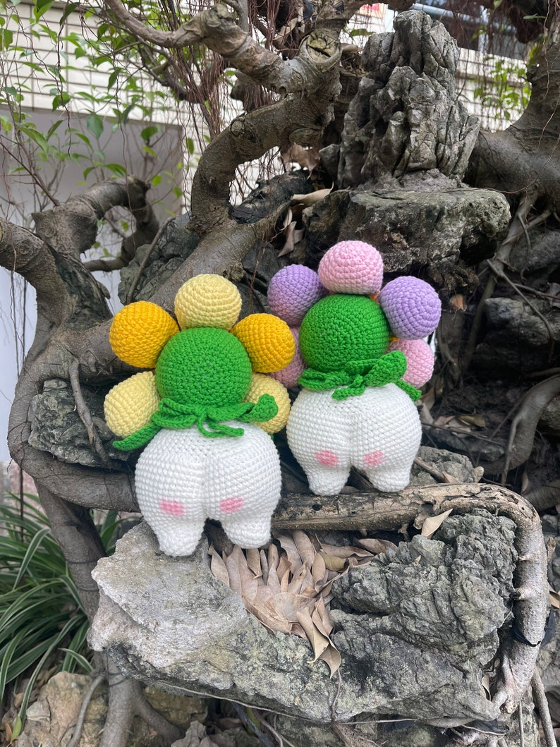 May include: Two crocheted plush toys shaped like flowers with white bodies and green leaves. The flowers have yellow, pink, and purple crocheted balls.
