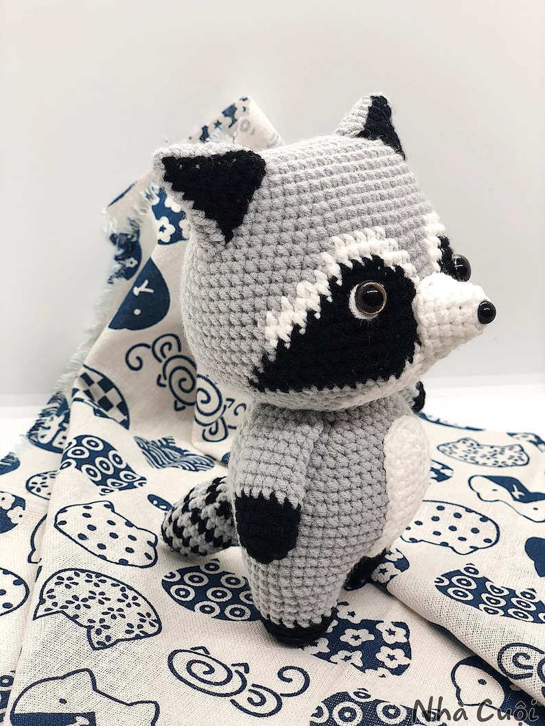 May include: A crocheted raccoon plush toy with black and white markings on its face and a black tail. The toy is standing on a white fabric with a blue and white pattern.