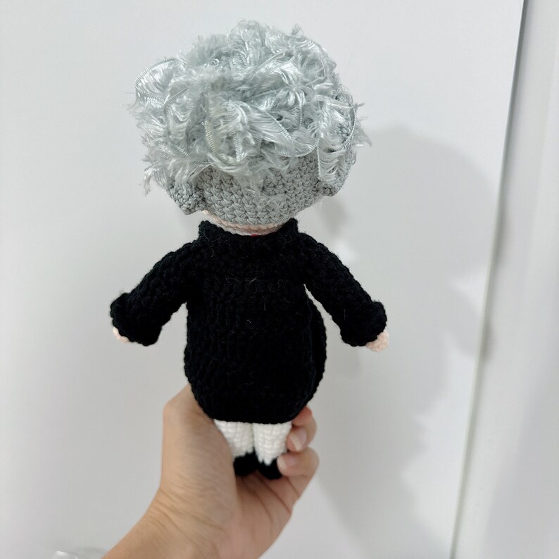 May include: A crocheted doll with gray curly hair, wearing a black sweater and white pants.