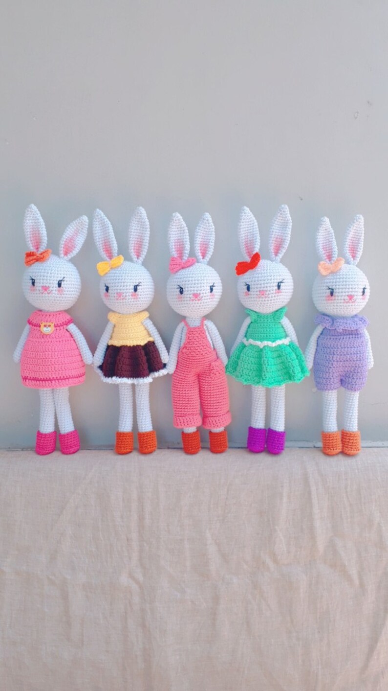May include: Five crocheted bunny dolls with different colored outfits. The bunnies are white with pink ears and black eyes. The outfits include pink, brown, green, and purple.