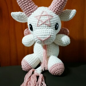 May include: A white crocheted goat with pink horns, hooves, and a tail. The goat has a pink pentagram on its forehead and black eyes.
