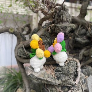May include: Two crocheted stuffed toys shaped like flowers. The first toy is yellow and brown with green leaves. The second toy is pink, purple, and white with green leaves. Both toys are sitting on a tree branch.