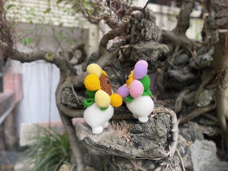 May include: Two crocheted stuffed toys shaped like flowers. The first toy is yellow and brown with green leaves. The second toy is pink, purple, and white with green leaves. Both toys are sitting on a tree branch.