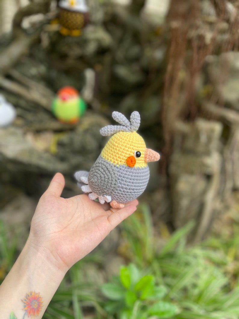 May include: A crocheted gray and yellow bird with a small orange beak and a red eye. The bird is being held in a person's hand.