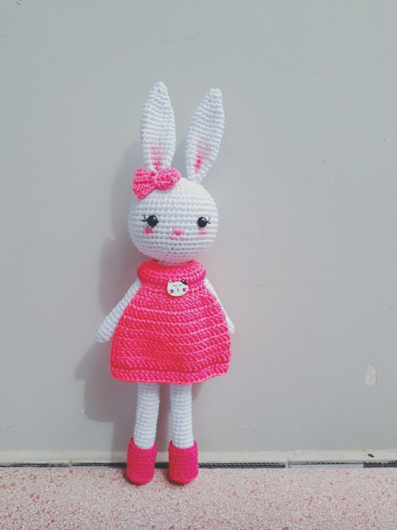 May include: A white crocheted bunny doll wearing a pink dress and pink shoes. The bunny has a pink bow on its head and a white cat button on its dress.