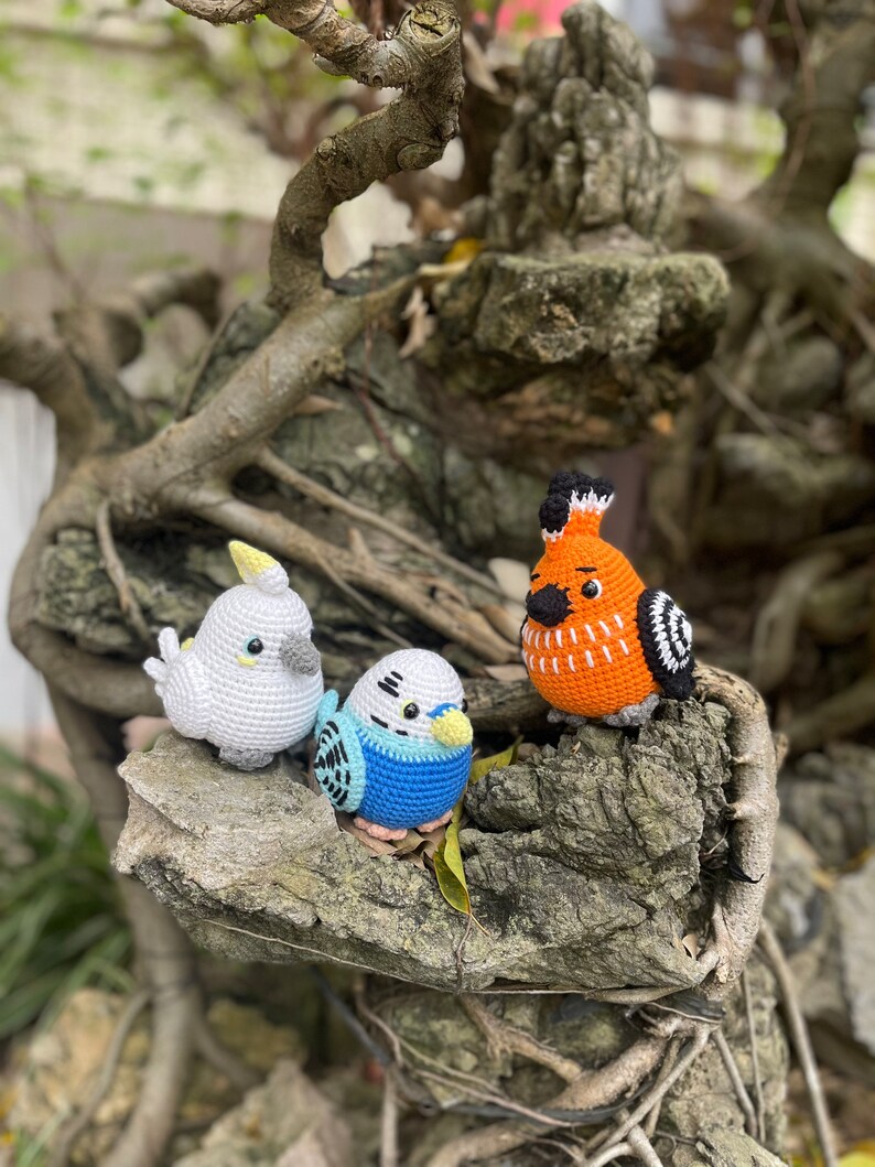 May include: Three crocheted bird toys, a white cockatiel, a blue and black budgie, and an orange and black cardinal, are perched on a tree branch.