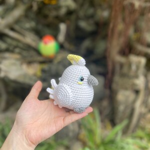 May include: A crocheted white cockatoo with a yellow crest and gray feet. The cockatoo is being held in a person's hand.