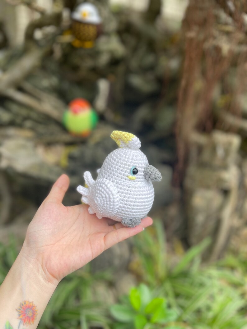 May include: A crocheted white cockatoo with a yellow crest and gray feet. The cockatoo is being held in a person's hand.