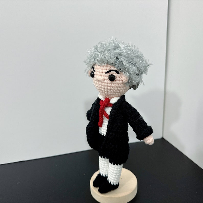 May include: A crocheted doll with gray hair, a black suit, a red tie, and white pants. The doll is standing on a wooden base.