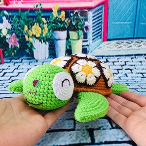 May include: A crocheted green turtle with a brown shell decorated with white and yellow flowers. The turtle is being held in a person's hands.