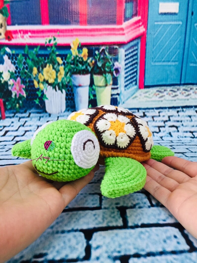 May include: A crocheted green turtle with a brown shell decorated with white and yellow flowers. The turtle is being held in a person's hands.