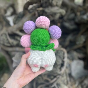 May include: A crocheted stuffed toy shaped like a flower with a green center and white body. The flower has pink and purple crocheted balls.