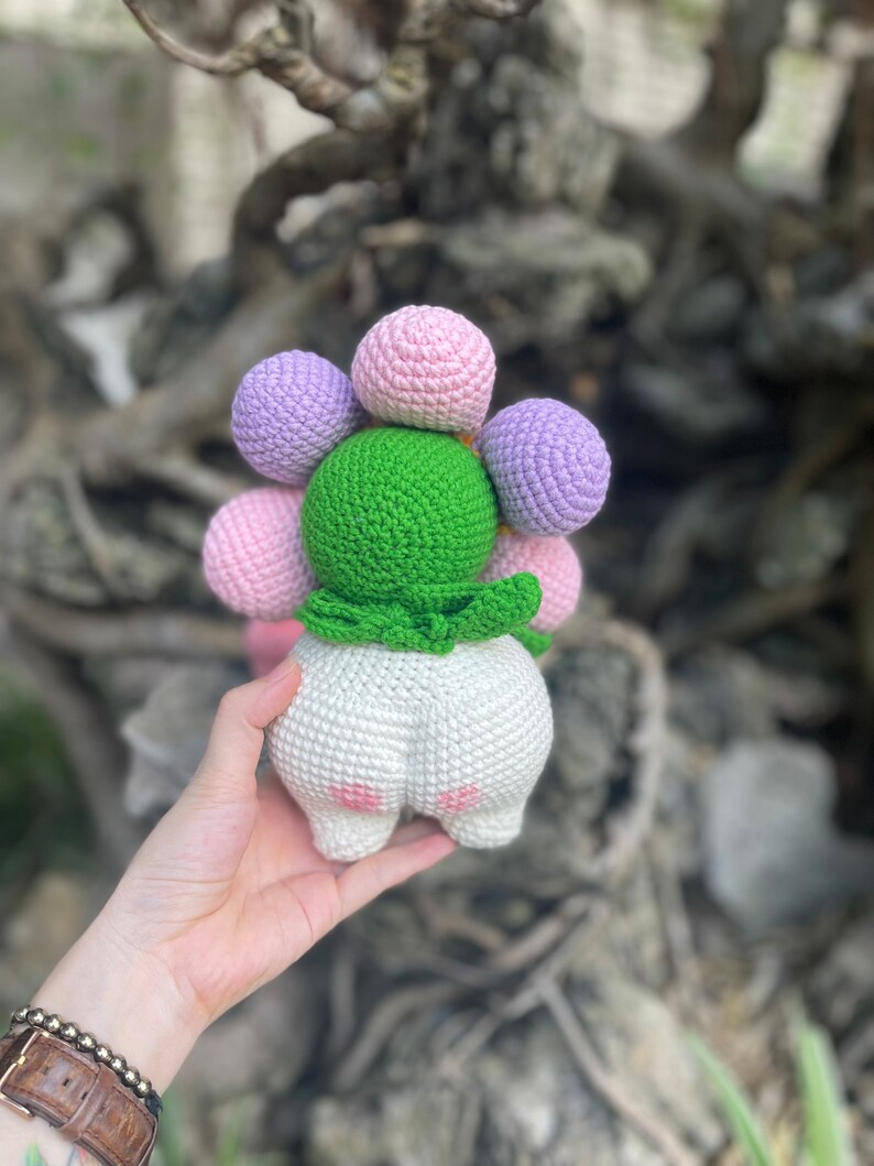 May include: A crocheted stuffed toy shaped like a flower with a green center and white body. The flower has pink and purple crocheted balls.