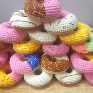 Op de afbeelding: Een stapel gehaakte donuts in verschillende kleuren en ontwerpen, waaronder roze, bruin, wit, geel en groen. De donuts zijn versierd met sprankelingen, glazuur en chocolade.