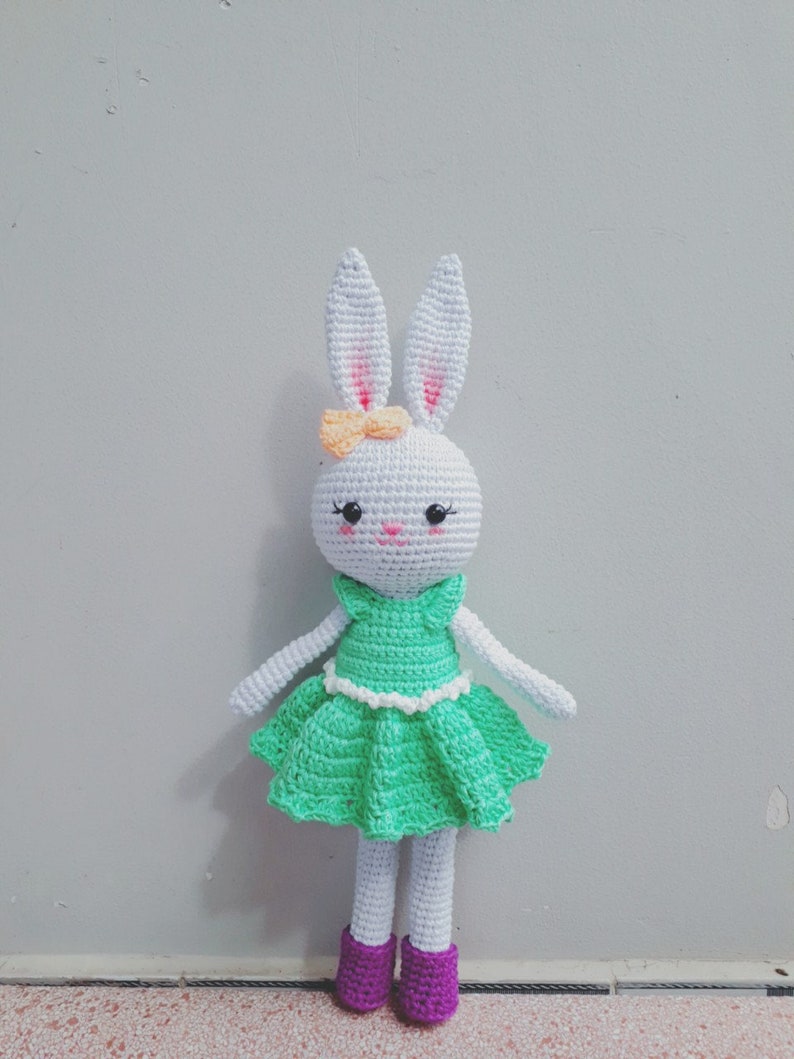 May include: A white crocheted bunny doll wearing a green dress with white trim and purple shoes. The bunny has a pink nose and orange bow.