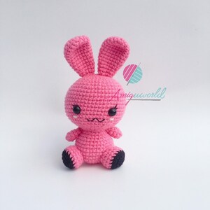 May include: A pink crocheted bunny rabbit with black eyes and a black nose. The bunny is sitting on a white surface.