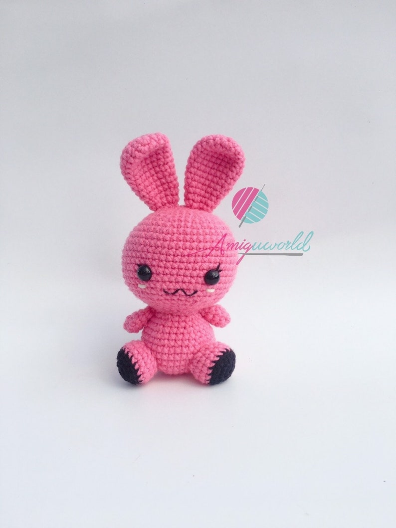 May include: A pink crocheted bunny rabbit with black eyes and a black nose. The bunny is sitting on a white surface.