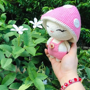 May include: A pink and white crocheted mushroom-shaped amigurumi doll with a white face and black eyes. The doll is wearing a pink crocheted dress with a button.