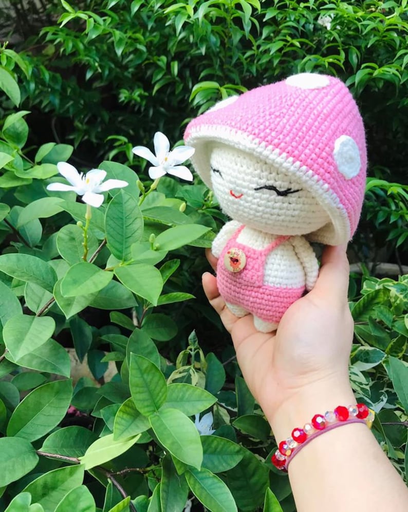 May include: A pink and white crocheted mushroom-shaped amigurumi doll with a white face and black eyes. The doll is wearing a pink crocheted dress with a button.