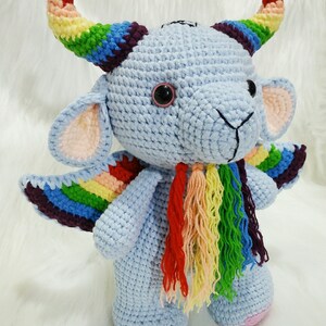 May include: A crocheted stuffed toy with a blue body, rainbow horns, wings, and tail. The toy has a cute, cartoonish face with black eyes and a pink nose.