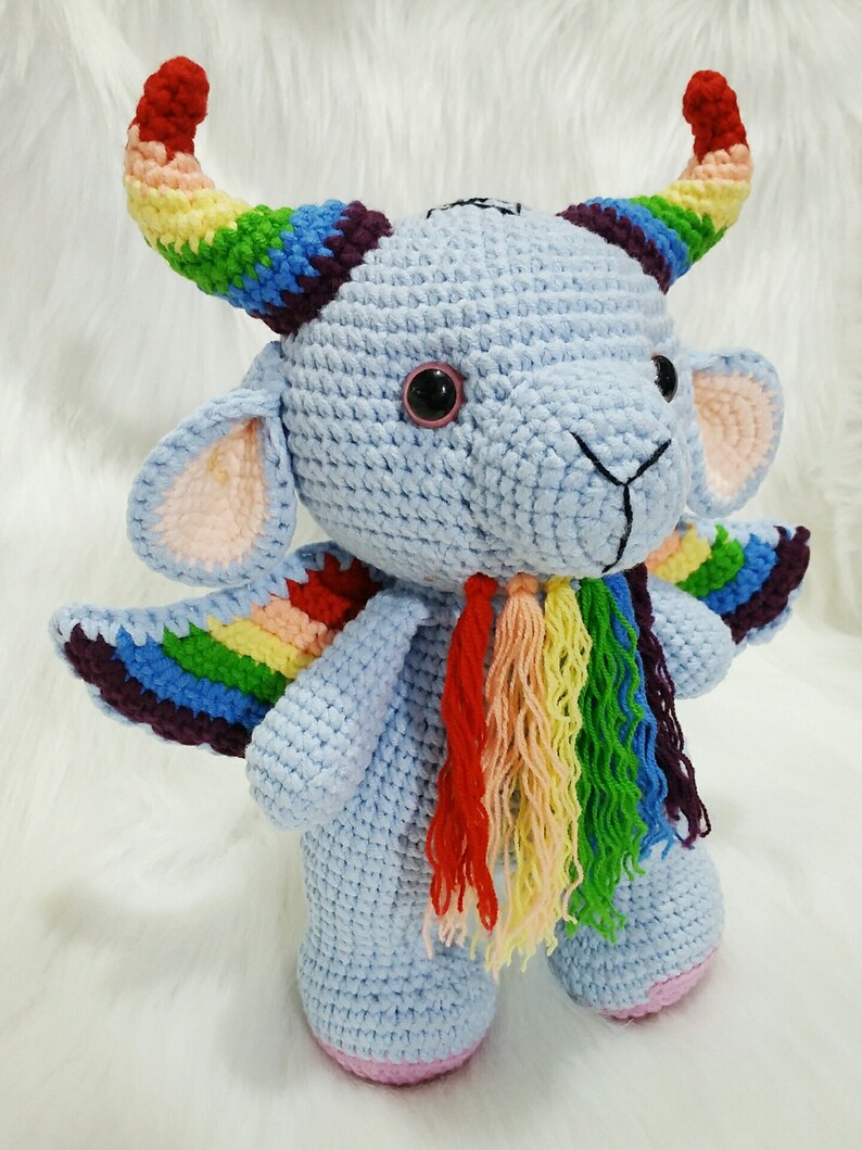 May include: A crocheted stuffed toy with a blue body, rainbow horns, wings, and tail. The toy has a cute, cartoonish face with black eyes and a pink nose.