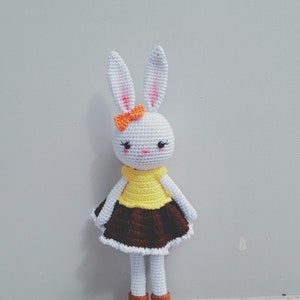 May include: A white crocheted bunny doll wearing a yellow and brown dress with an orange bow. The doll has black eyes and a pink nose.