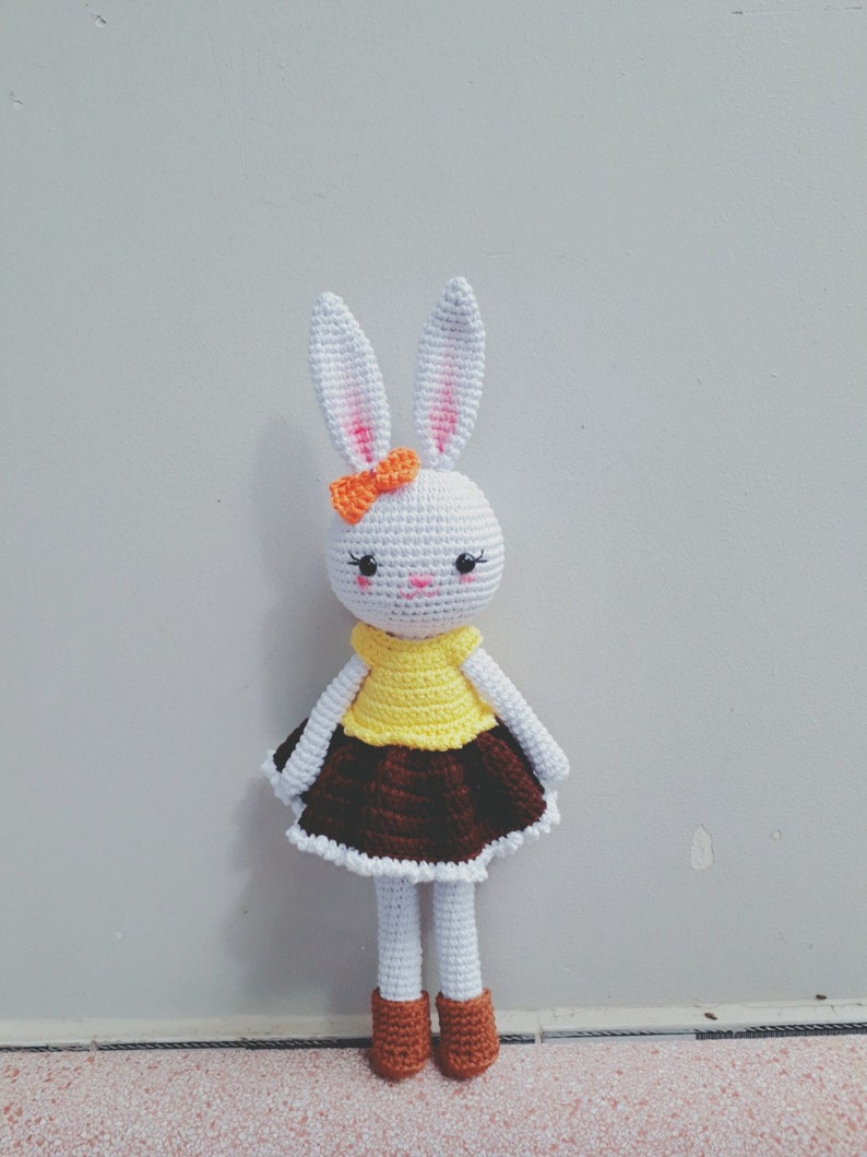 May include: A white crocheted bunny doll wearing a yellow and brown dress with an orange bow. The doll has black eyes and a pink nose.