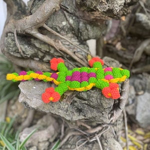 May include: A crocheted lizard toy with a green, yellow, pink, and orange color scheme. The toy is lying on a rock with a brown and gray bark-like texture.