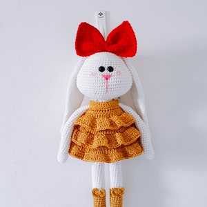 May include: A white crocheted bunny doll wearing a yellow ruffled dress and a red bow. The doll has black eyes and a pink nose.