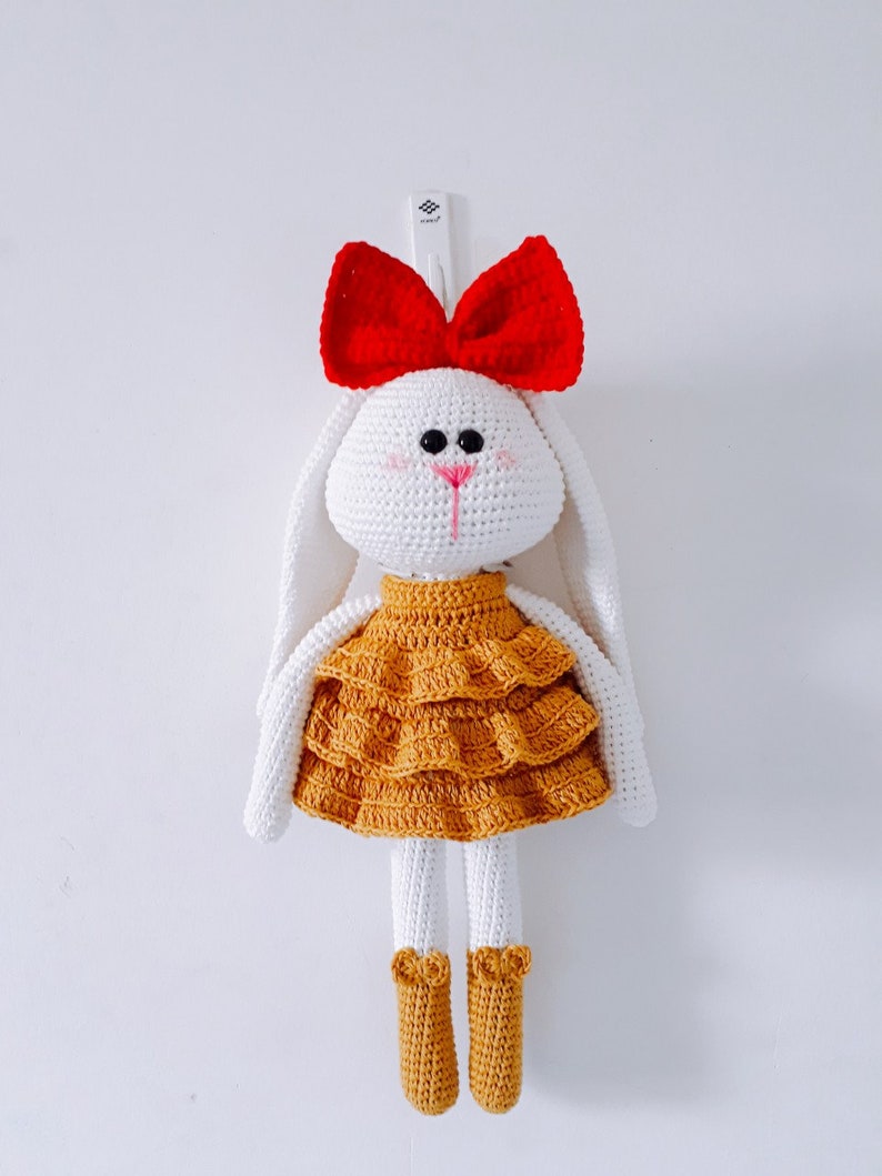 May include: A white crocheted bunny doll wearing a yellow ruffled dress and a red bow. The doll has black eyes and a pink nose.
