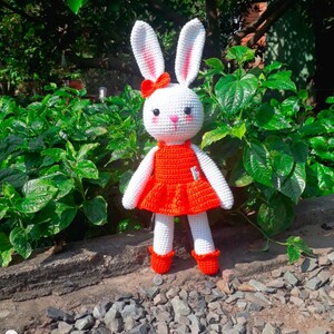 May include: White crocheted bunny wearing a red dress with a white bow. The bunny is standing on a bed of small rocks.