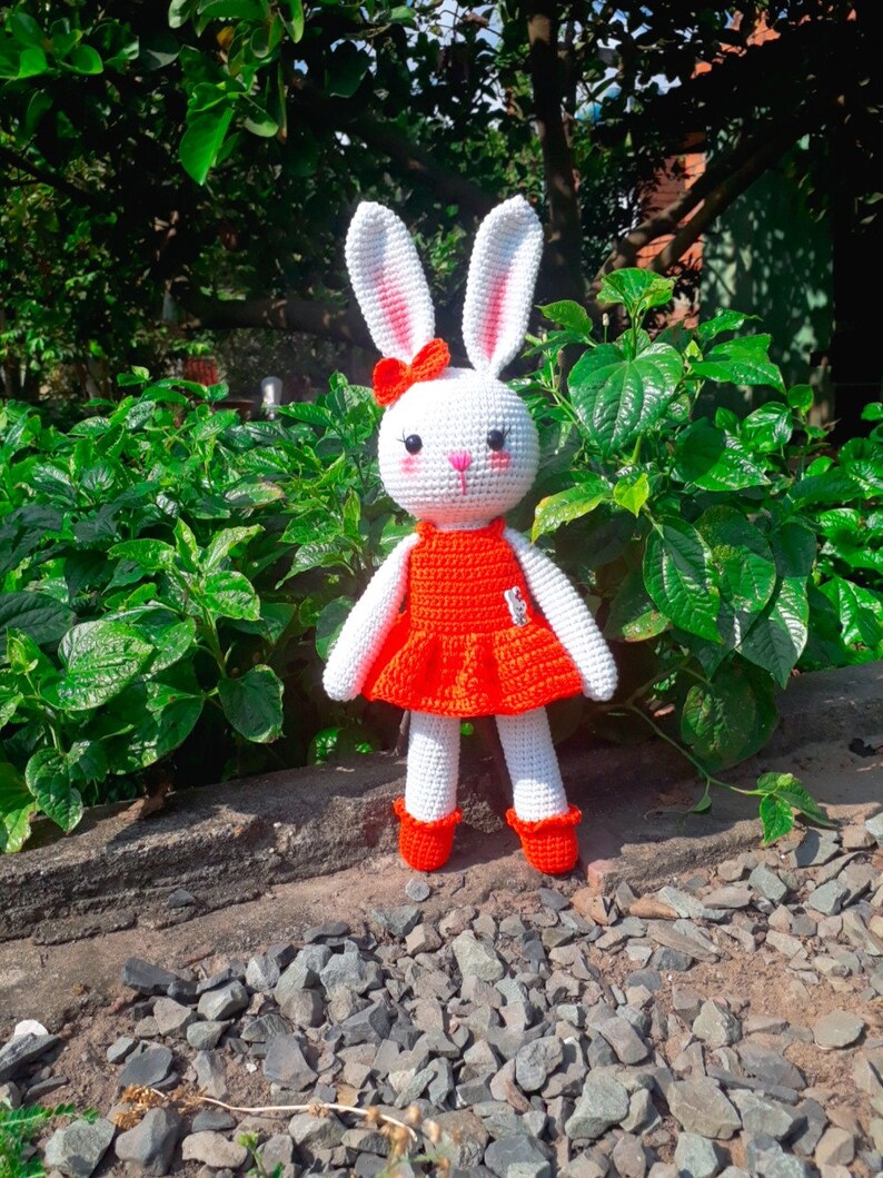 May include: White crocheted bunny wearing a red dress with a white bow. The bunny is standing on a bed of small rocks.
