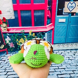 May include: A crocheted green turtle with a brown shell and white eyes. The shell is decorated with yellow and white flowers. The turtle is being held in a hand.