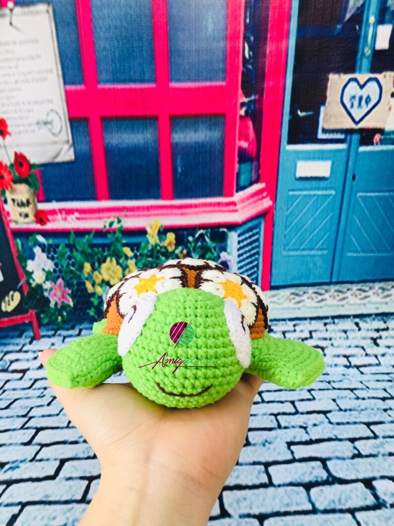 May include: A crocheted green turtle with a brown shell and white eyes. The shell is decorated with yellow and white flowers. The turtle is being held in a hand.
