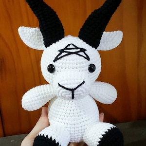 May include: White crocheted goat plush toy with black horns, eyes, and hooves. The goat has a black pentagram on its forehead.