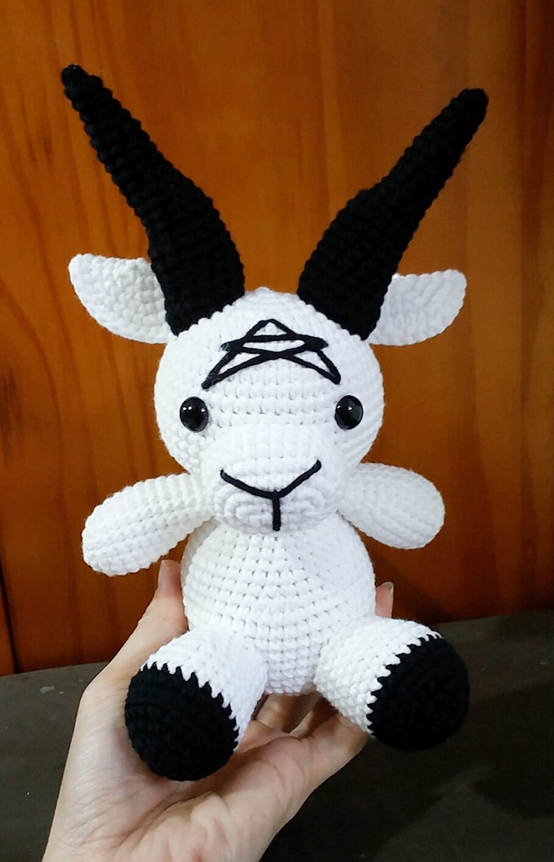 May include: White crocheted goat plush toy with black horns, eyes, and hooves. The goat has a black pentagram on its forehead.