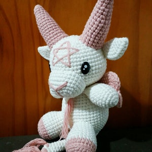 May include: A white crocheted goat with pink horns, hooves, and a tail. The goat has black eyes and a pink stitched design on its face.