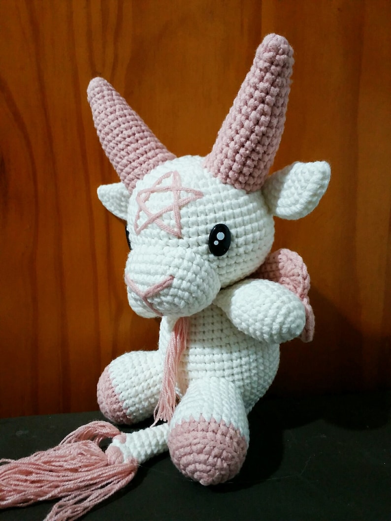May include: A white crocheted goat with pink horns, hooves, and a tail. The goat has black eyes and a pink stitched design on its face.