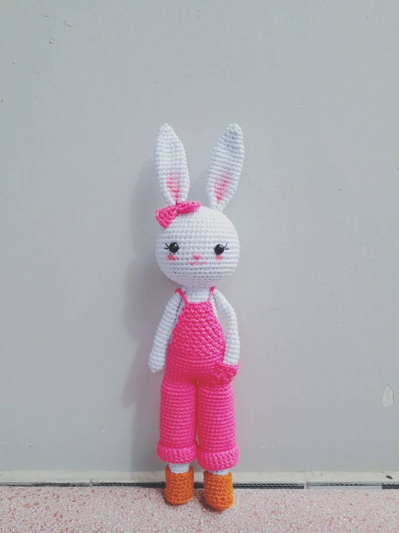 May include: A white crocheted bunny wearing pink overalls and orange shoes. The bunny has black eyes and a pink bow.