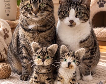 May include: Two adult tabby cats sitting side by side, with two small, handmade crocheted tabby cat figurines positioned in front of them. The figurines feature green eyes and detailed brown and white yarn patterns, mirroring the cats' markings.