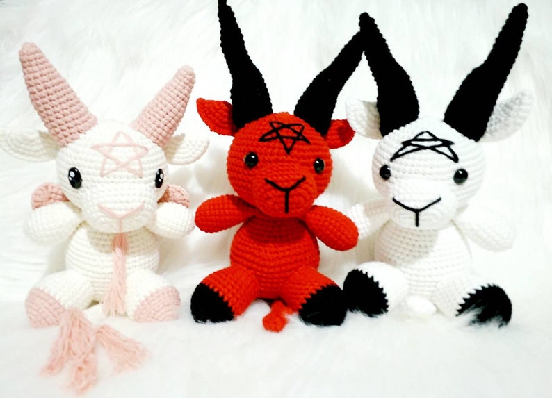 May include: Three crocheted goat plushies, one red, one white with pink accents, and one white with black accents. Each plush has a pentagram on its forehead and horns.