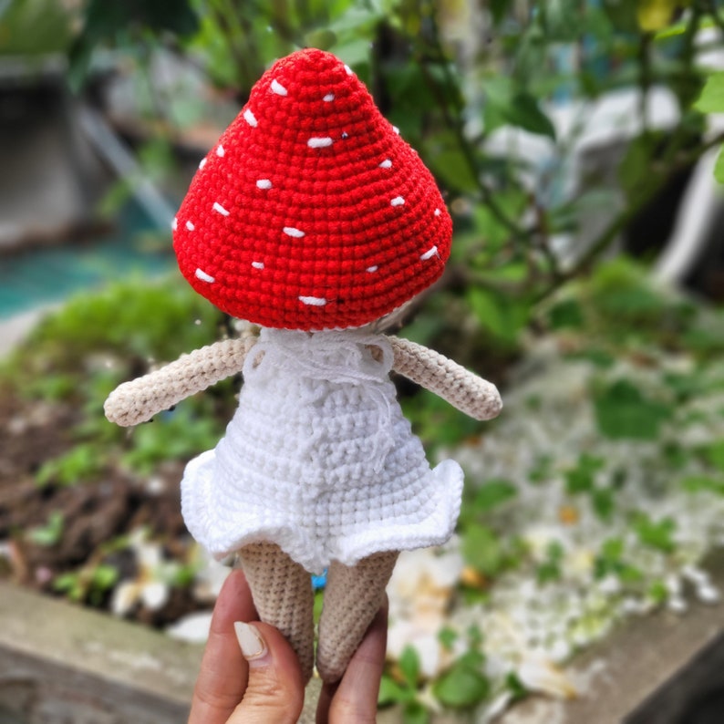 May include: A crocheted doll wearing a red and white mushroom hat and a white dress. The doll has a white crocheted dress with a ruffled hem.