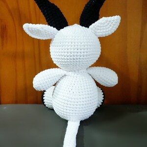May include: White crocheted goat with black horns and a long tail. The goat is sitting on a gray surface.