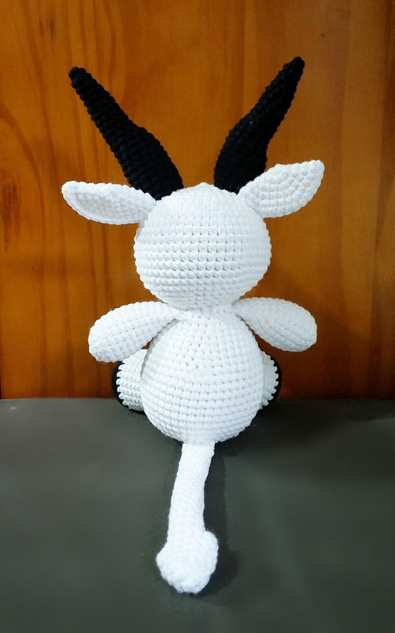 May include: White crocheted goat with black horns and a long tail. The goat is sitting on a gray surface.