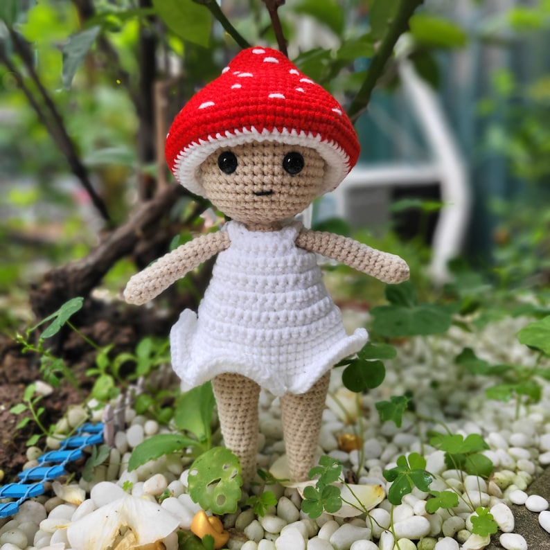 May include: A crocheted doll wearing a red and white mushroom hat and a white dress. The doll is standing on a bed of white stones.