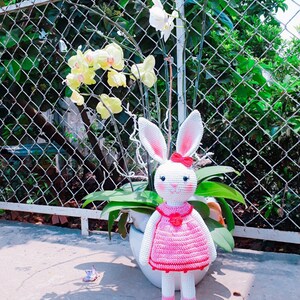 May include: A white crocheted bunny wearing a pink dress with red trim and a red flower on the chest. The bunny is standing on a white stand in front of a potted plant with white and yellow orchids.