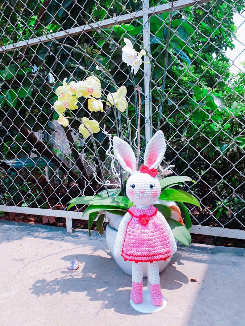 May include: A white crocheted bunny wearing a pink dress with red trim and a red flower on the chest. The bunny is standing on a white stand in front of a potted plant with white and yellow orchids.