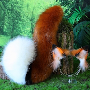 Puede incluir: Un conjunto de accesorios de cosplay con temática de zorro, que incluye una cola roja-naranja esponjosa, una cola blanca y negra y una diadema con orejas de zorro. Las orejas son naranjas, blancas y negras. Los artículos se exhiben sobre musgo.