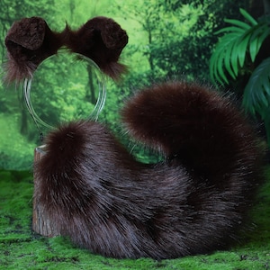 以下が含まれることがあります： 茶色のフェイクファーの動物の耳のヘッドバンドと尻尾のセット。耳は透明なヘッドバンドに取り付けられています。尻尾は大きく、ふわふわした、湾曲した形をしています。セットは、緑色の苔むした表面に、森の背景で表示されています。